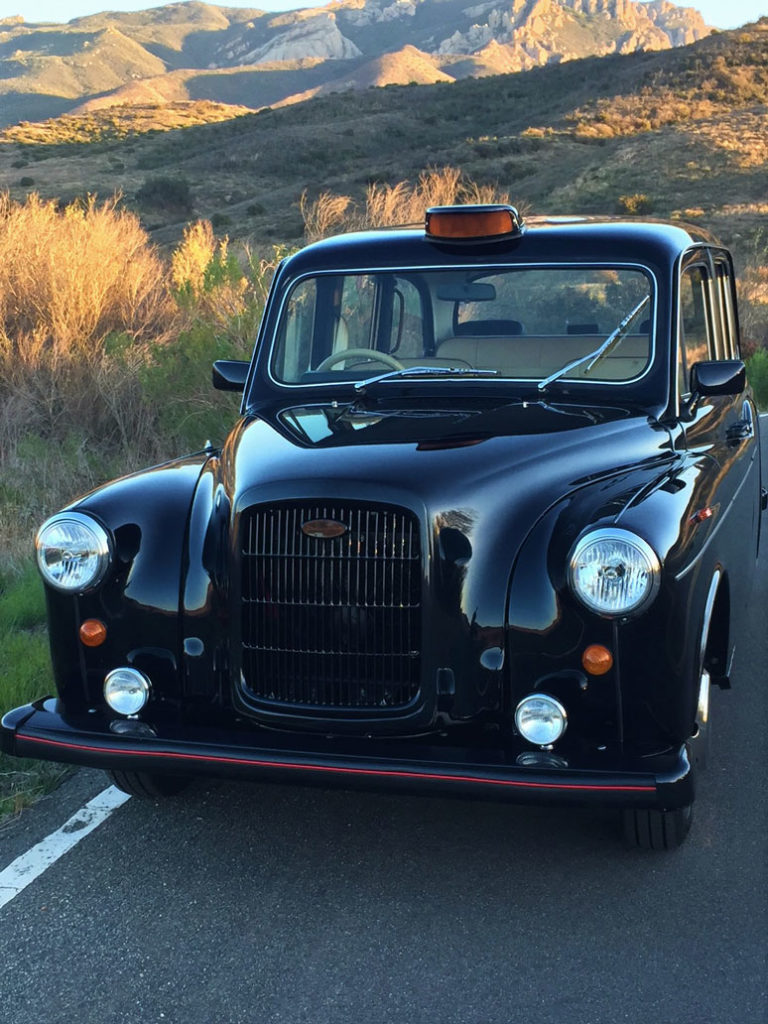Front view of dark colored London Taxi against a hilly backdrop